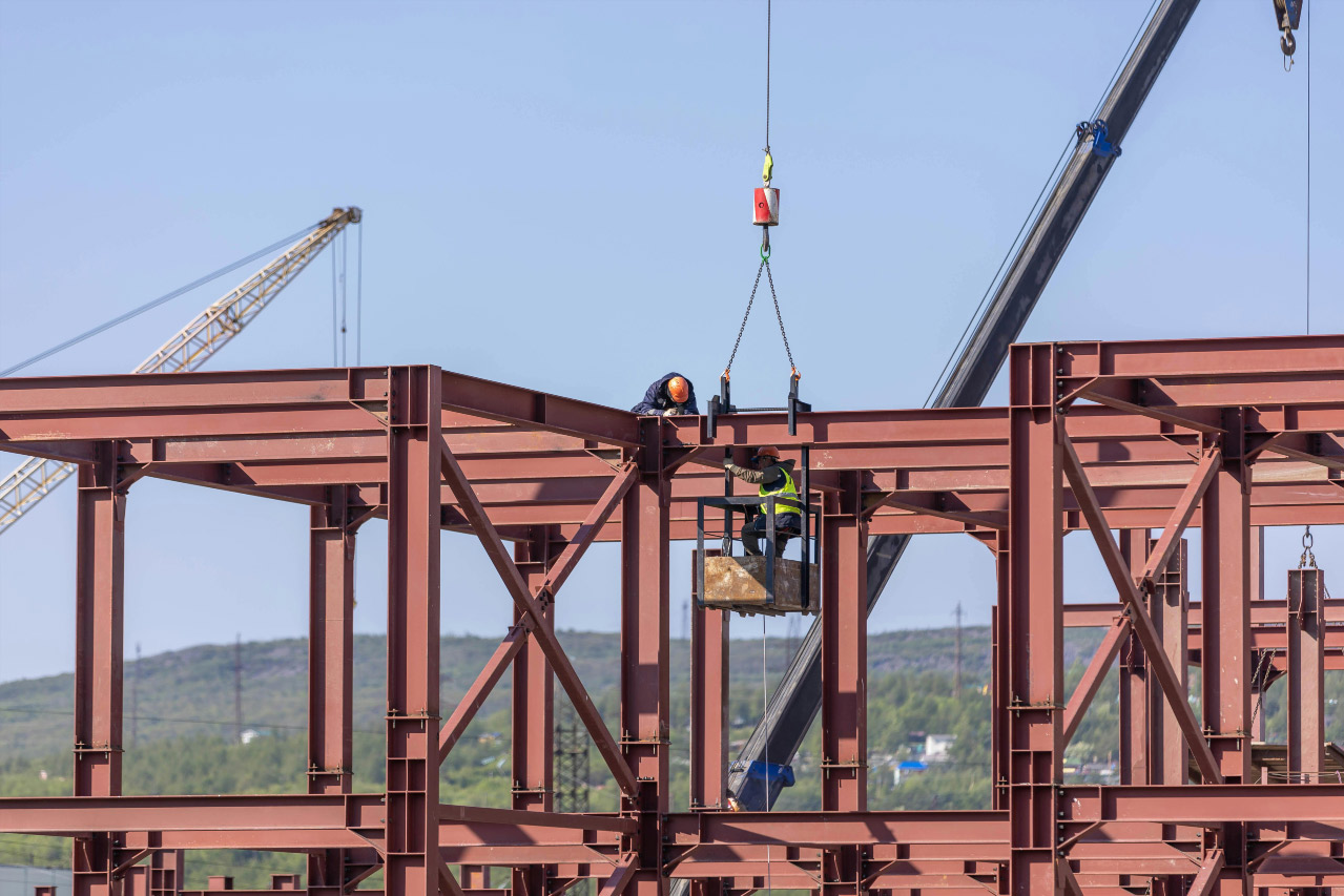 Estrutura metálica em montagem com vigas de aço, profissionais de obra e equipamentos de segurança.